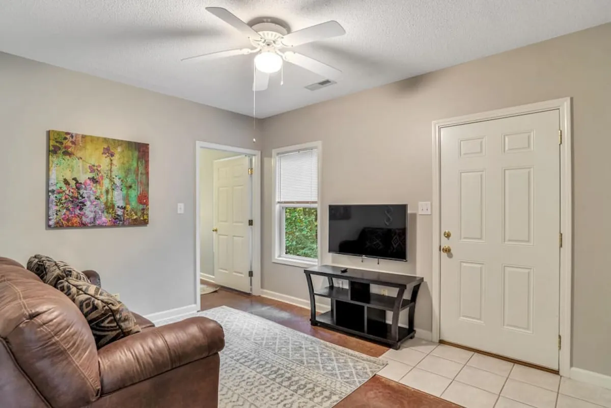 This image depicts a comfortable and inviting living room anchored by a leather sofa, artwork on the wall, a television unit, and a window that lets in natural light. The scene suggests a communal space ideal for gathering, relaxing, and planning family activities after a day exploring Asheville.