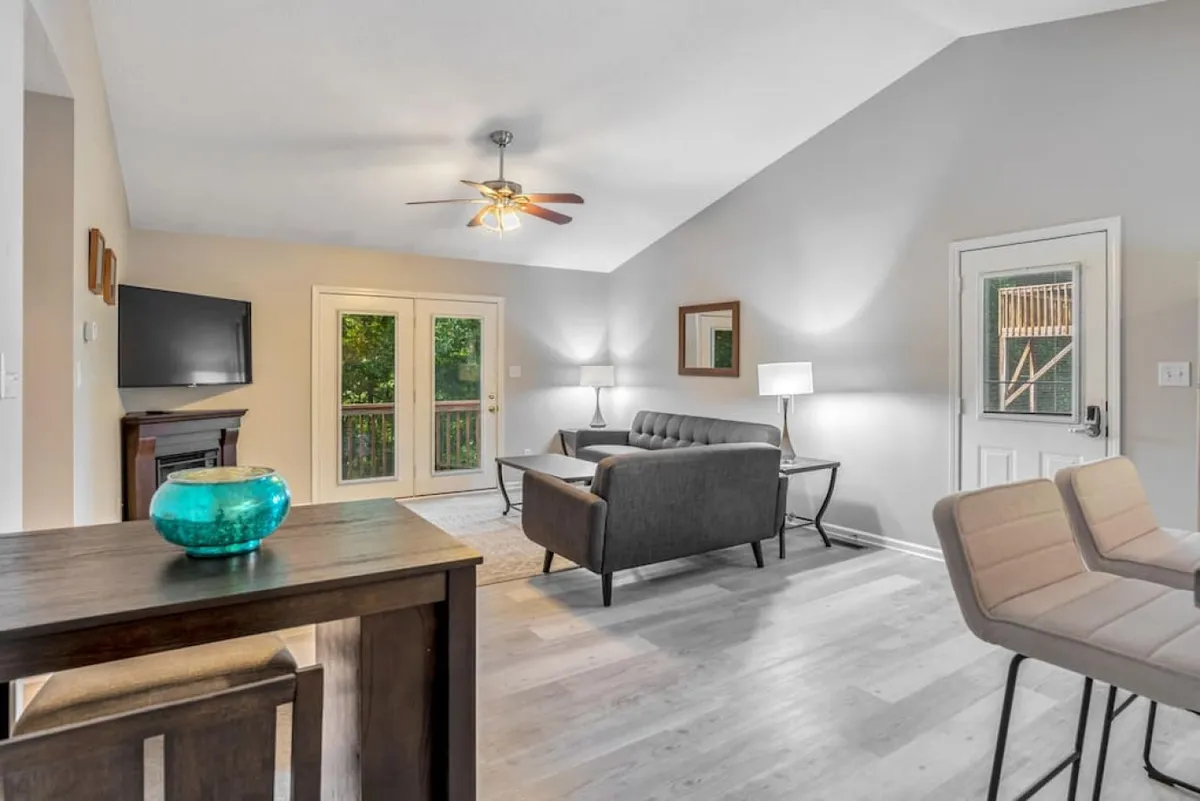 A cozy, sunlit living and dining area featuring a sofa, coffee table, TV, and bar stools at the kitchen counter. Glass doors open to an outdoor space, and the layout encourages socializing, relaxing, and casual meals. The arrangement allows for seamless interaction between the kitchen, dining, and living spaces.