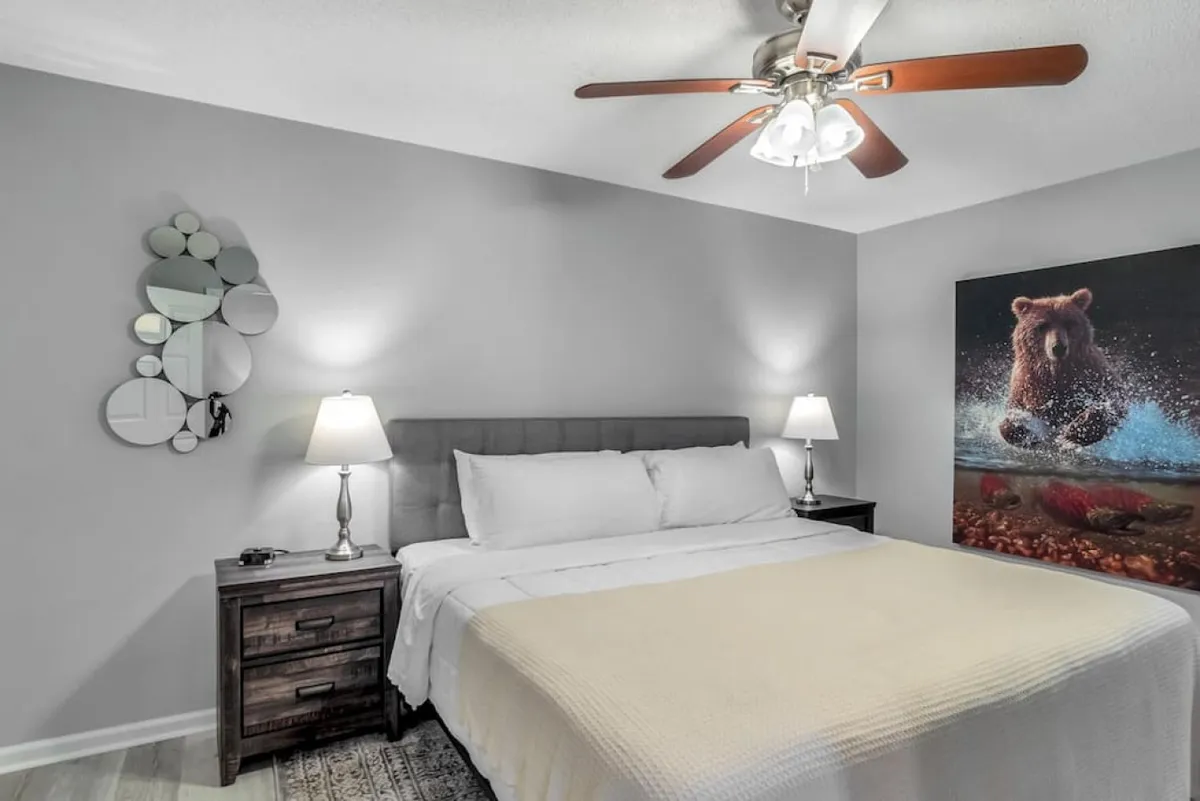 A peaceful bedroom scene with a bed dressed in plush white linens, a distinctive bear painting above the headboard, and a nightstand with a warm lamp glow. The space feels personal and serene, perfect for unwinding after a day of adventure.