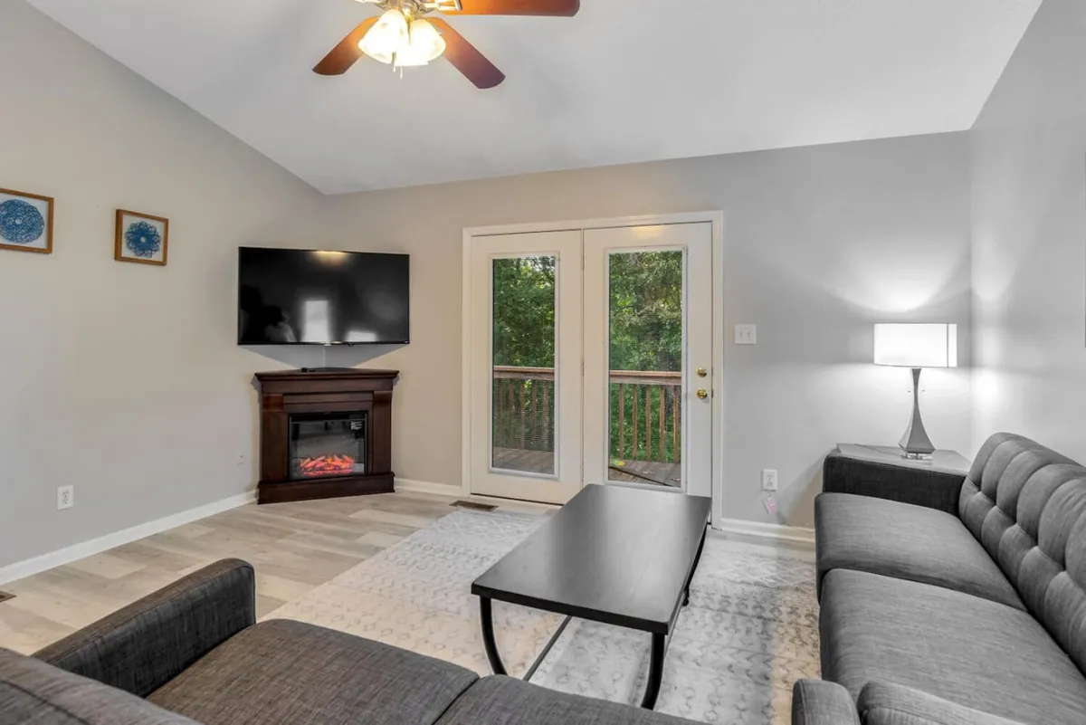 A welcoming living room anchored by a comfortable sofa faces a TV and a modern fireplace. Double doors open to a balcony, allowing abundant natural light to stream in and visually extending the open-concept living area. The space is styled with chic, contemporary furnishings and offers both relaxation and entertainment possibilities.