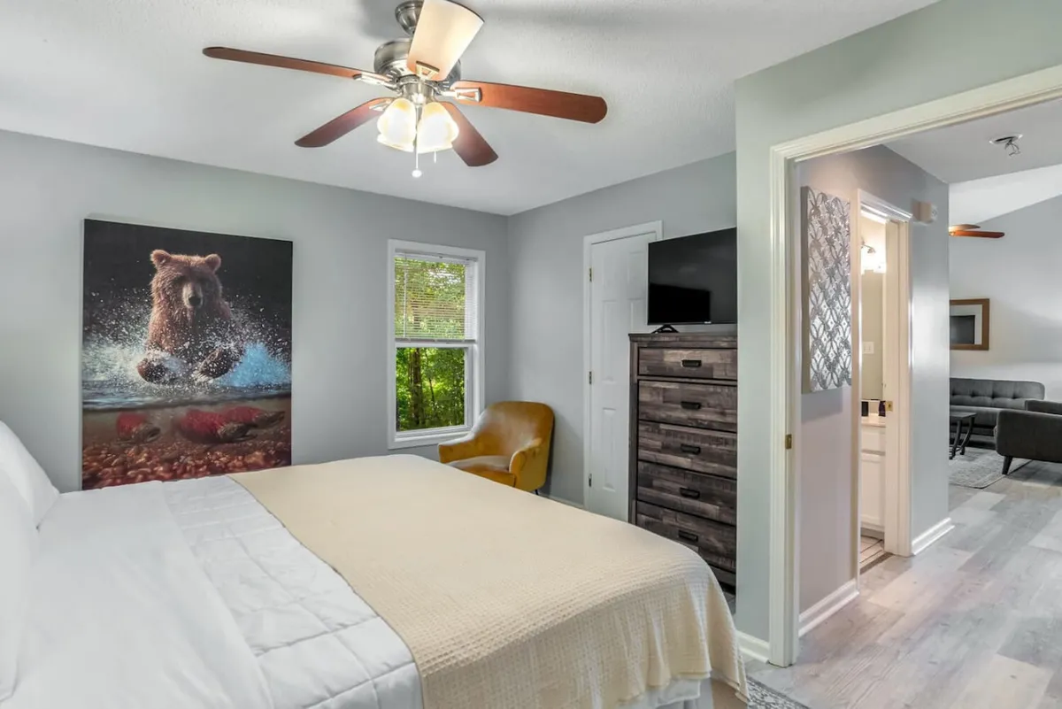 A cozy bedroom featuring a bed with crisp white bedding, a bear painting that nods to local wildlife, a wooden dresser, and a comfortable armchair. The space feels serene—a restful retreat after a day of hiking or exploring.