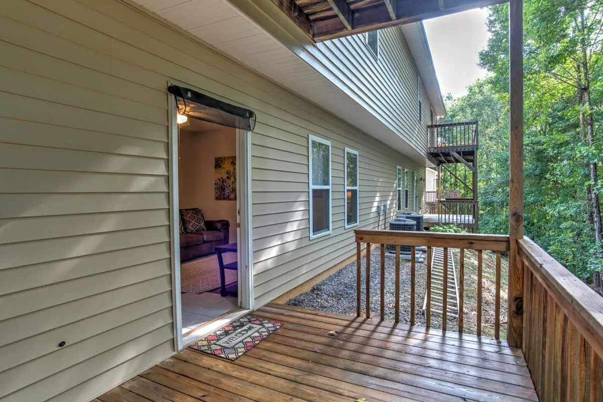 A wooden deck with an open door leading inside, surrounded by vibrant greenery. The space feels welcoming and seamlessly connected to the home's interior, suggesting easy movement between indoor and outdoor areas. Ample sunlight and inviting outdoor seating hint at opportunities for socializing or quiet relaxation.